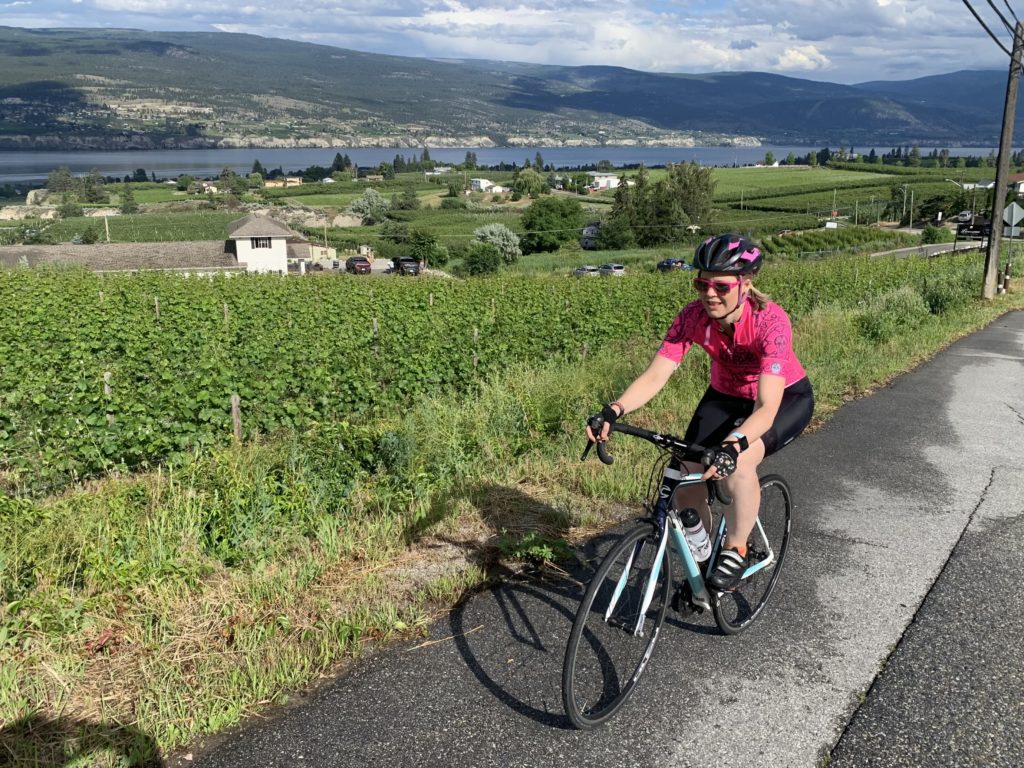 A female cyclist pedals up a steep hill with a vineyard next to her
