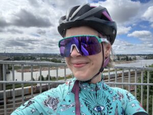 Female cyclist wheres new Bianchi Blue Oakley sunglasses with Fraser River and white clouds with blue sky behind