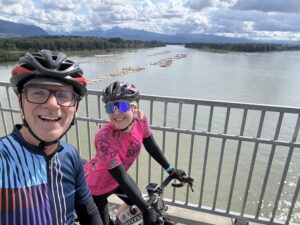 A female and male cyclist take a picture on the Port Mann Bridge with the Fraser River behind them.