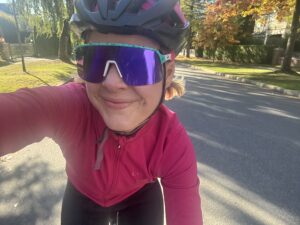 A female cyclist in a pink long-sleeve jersey takes a selfie while riding her bike. Trees with orange and yellow leaves in the background